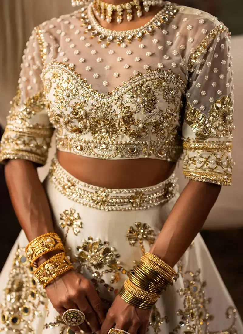 Ivory lehenga choli close-up showing a sheer pearl-dotted neckline, gold embroidered sleeves and mirrored waistband